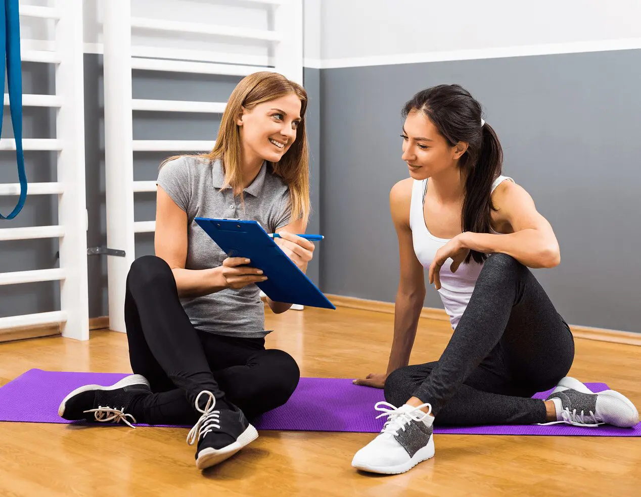 Two women sit on exercise mats in a gym, engaged in personalized fitness coaching. One holds a clipboard and pen, guiding the session, while the other listens attentively. Their discussion is focused and purposeful.