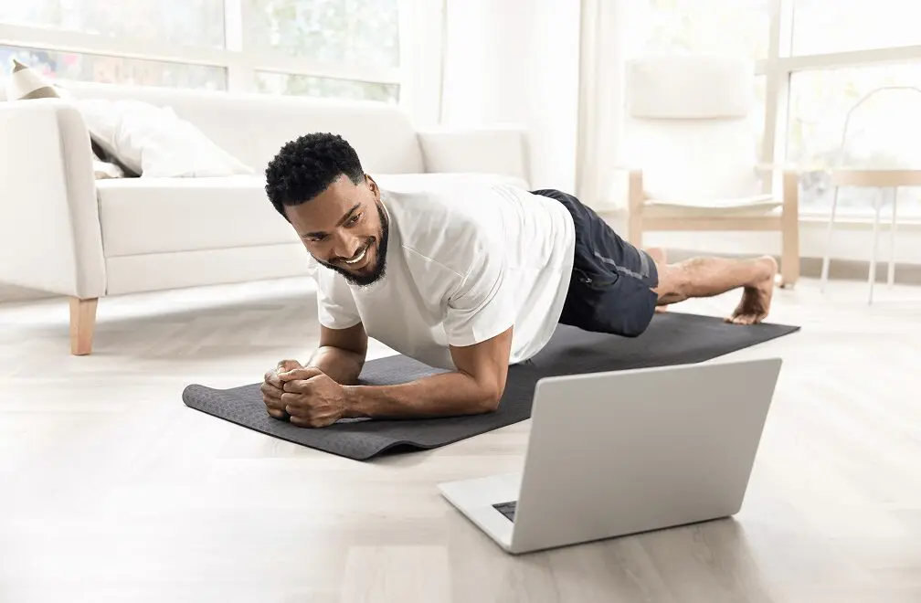A person engaged in a plank exercise on a yoga mat indoors, utilizing in-home personal training via a laptop screen.